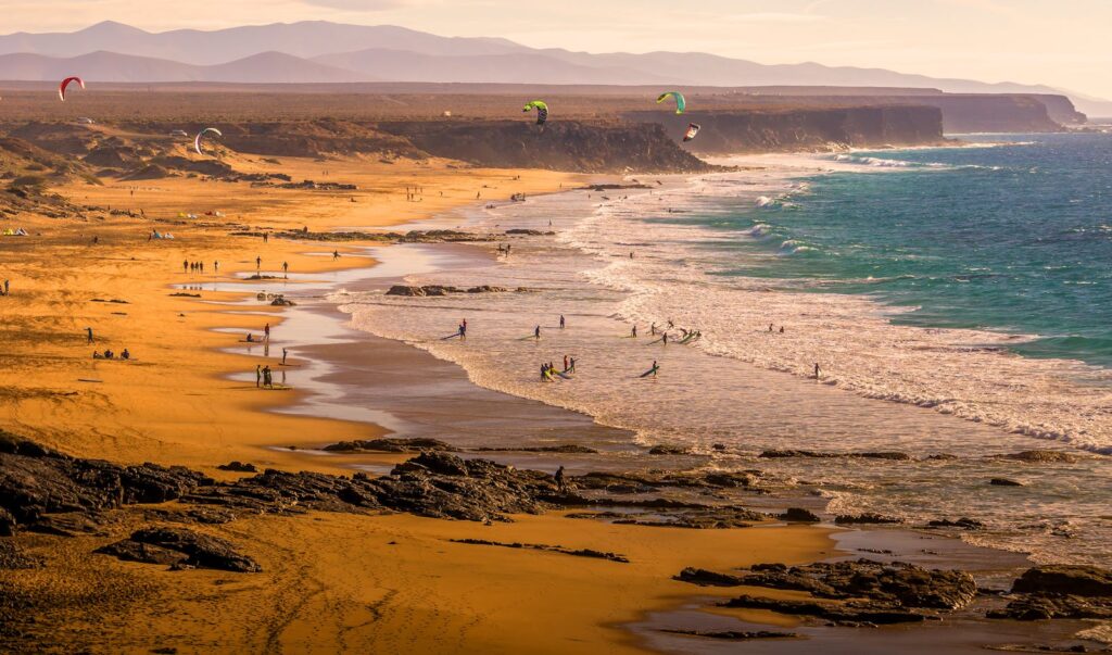 Enjoy a lively beach with kitesurfers and scenic cliffs at sunset, perfect for vacation wallpaper.