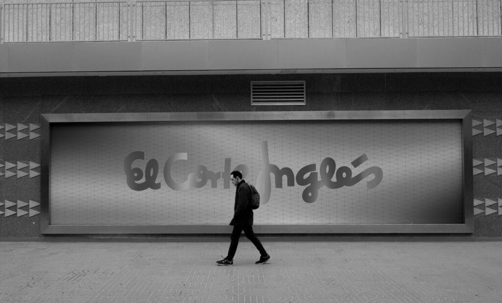 A man walks past El Corte Inglés sign in a monochrome city environment.